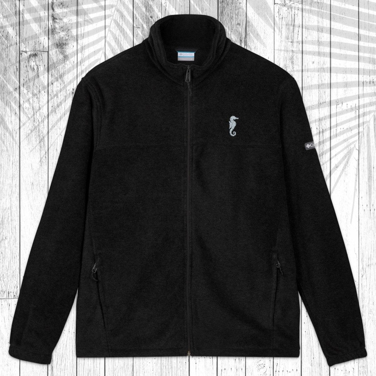 A black fleece zip-up jacket with a stand collar, shown flat against a white wood plank background with a palm leaf pattern overlay. The jacket features a small embroidered seahorse on the left chest and a small white logo on the right sleeve.