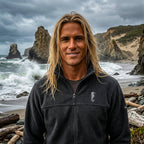 A man wearing a dark charcoal gray zip-up fleece jacket and khaki pants stands on a sandy beach path with the ocean and sky in the background.
