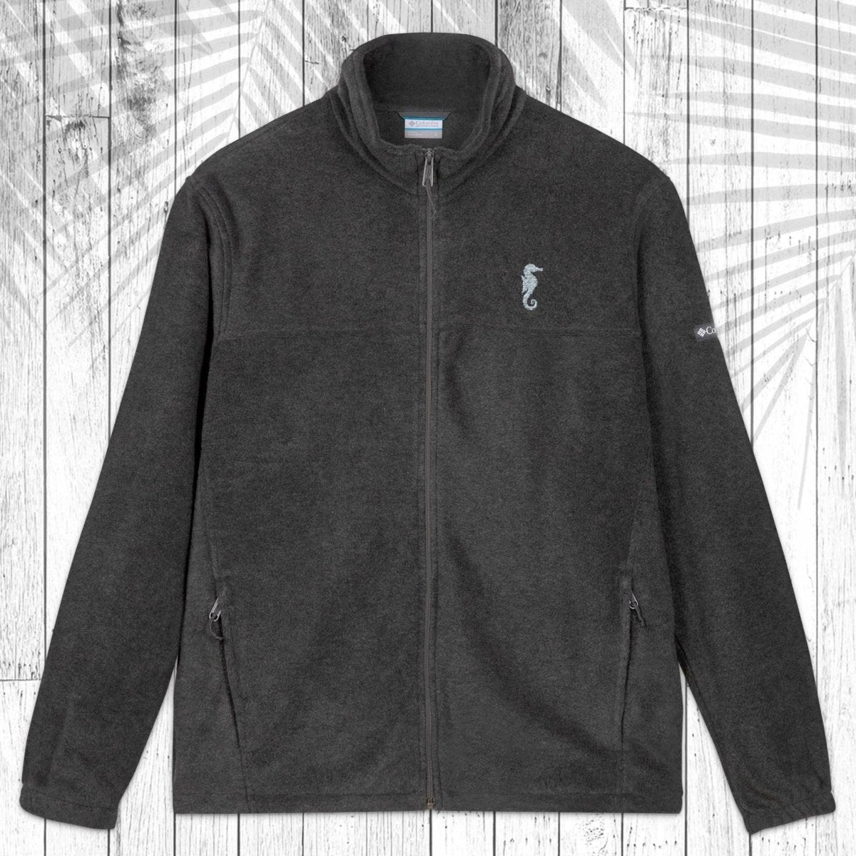 A dark gray fleece zip-up jacket with a stand collar, shown flat against a white wood plank background with a palm leaf shadow. The jacket features a small embroidered seahorse on the left chest and a small white logo on the right sleeve.
