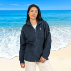 A woman stands on a beach wearing a dark navy fleece zip-up jacket with a white embroidered seahorse on the left chest, paired with light beige pants.