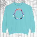 A light mint crewneck sweatshirt featuring a colorful graphic of an open mouth with sharp teeth on the chest, displayed against a white wood plank background with a palm leaf overlay.
