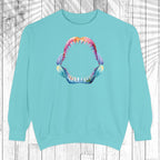 A light mint crewneck sweatshirt featuring a colorful graphic of an open mouth with sharp teeth on the chest, displayed against a white wood plank background with a palm leaf overlay.