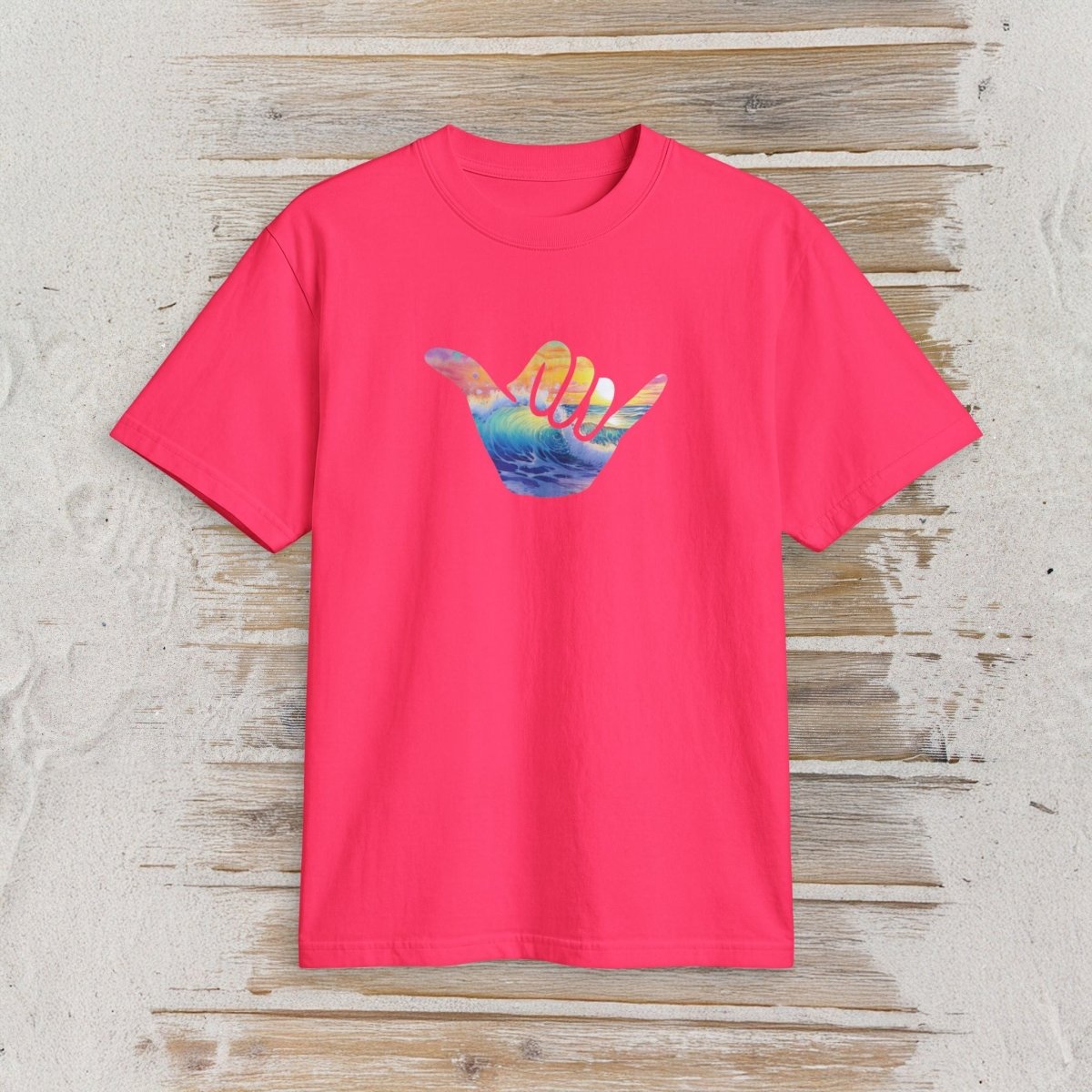 A bright pink short-sleeve t-shirt with a colorful graphic of a hand in a shaka sign, featuring a sunset landscape inside the hand. The t-shirt is displayed flat on a light-colored, rustic wooden plank background.