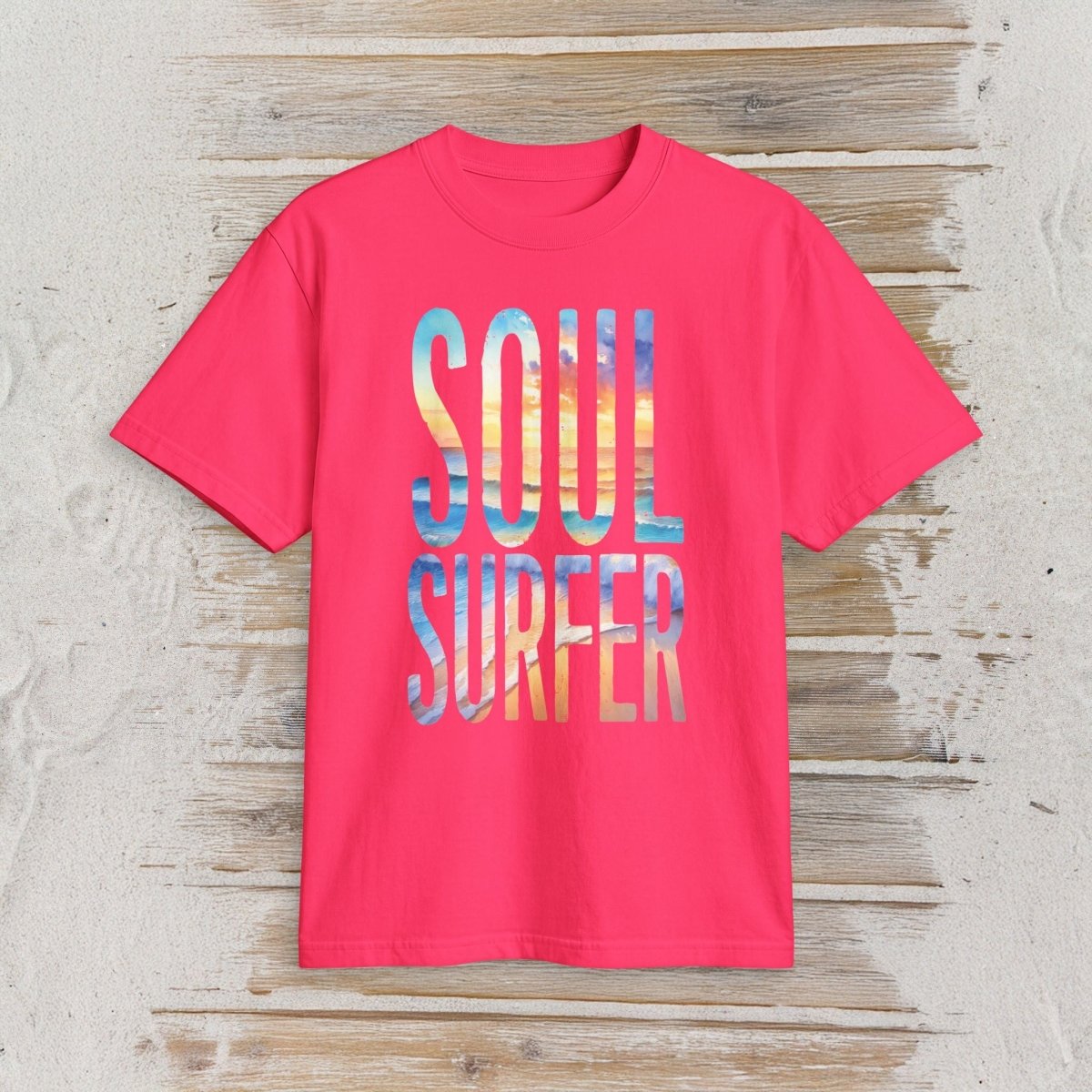 A bright pink short-sleeve t-shirt with a colorful graphic reading 'SOUL SURFER' in a watercolor-style beach scene of blue, yellow, and orange, laid flat on a rustic light wood background.