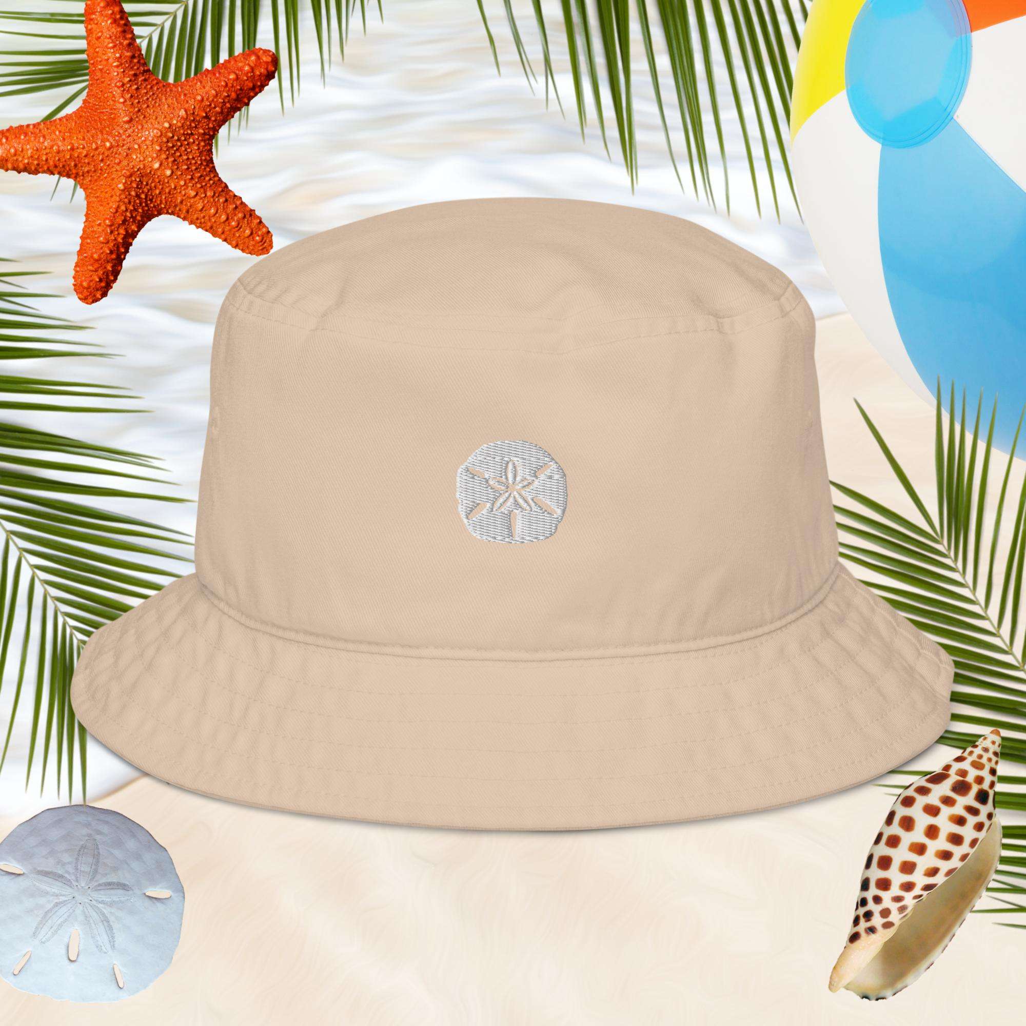Beige cotton bucket hat with an embroidered sand dollar design on a beach-themed background with palm leaves, starfish, and shells.