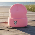 A light pink knit beanie hat with a folded cuff, featuring a small embroidered shark tooth graphic in black and white on the front.