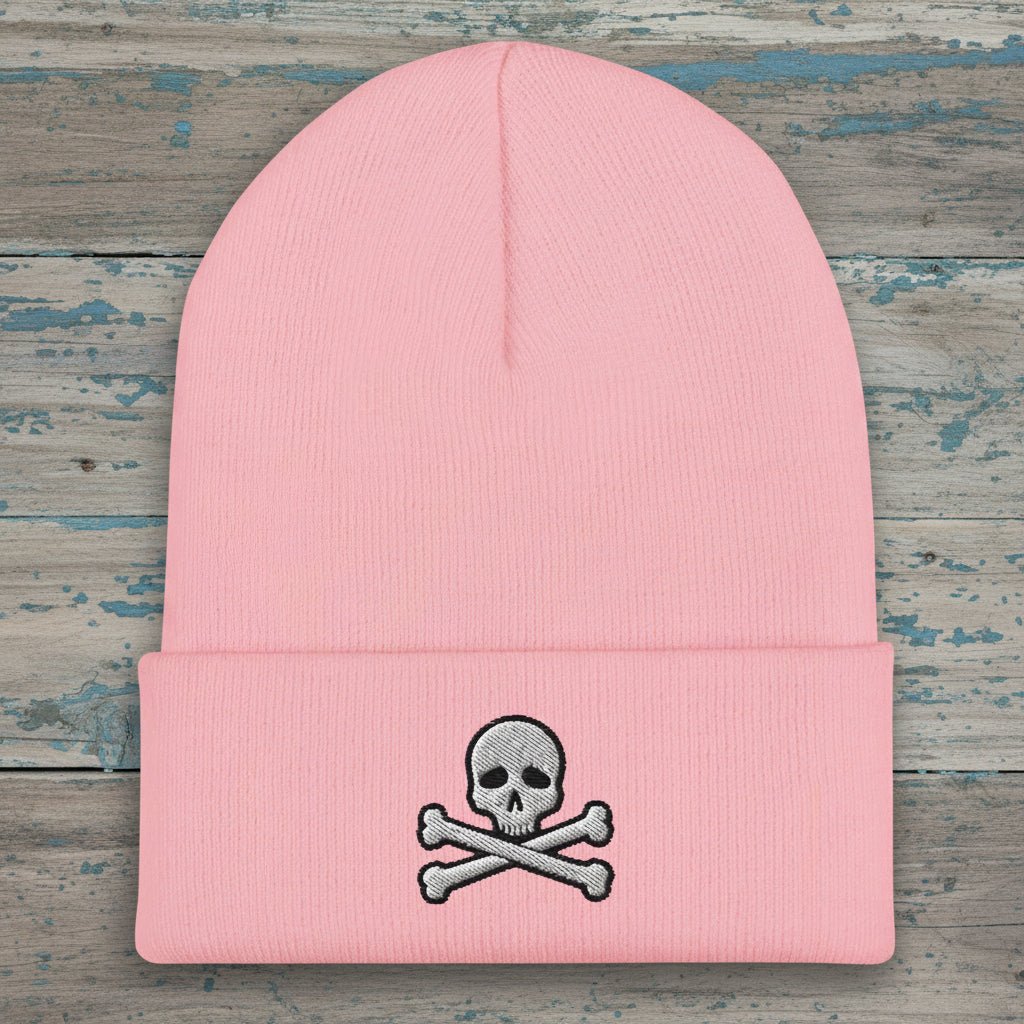 A light pink ribbed knit beanie hat with a folded cuff, featuring a small embroidered gray and white skull and crossbones on the front.