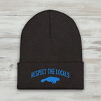 A dark charcoal gray heather knit beanie with a folded cuff, featuring blue embroidered text reading 'RESPECT THE LOCALS' and a blue manatee graphic on the front, set against a rustic wood background.
