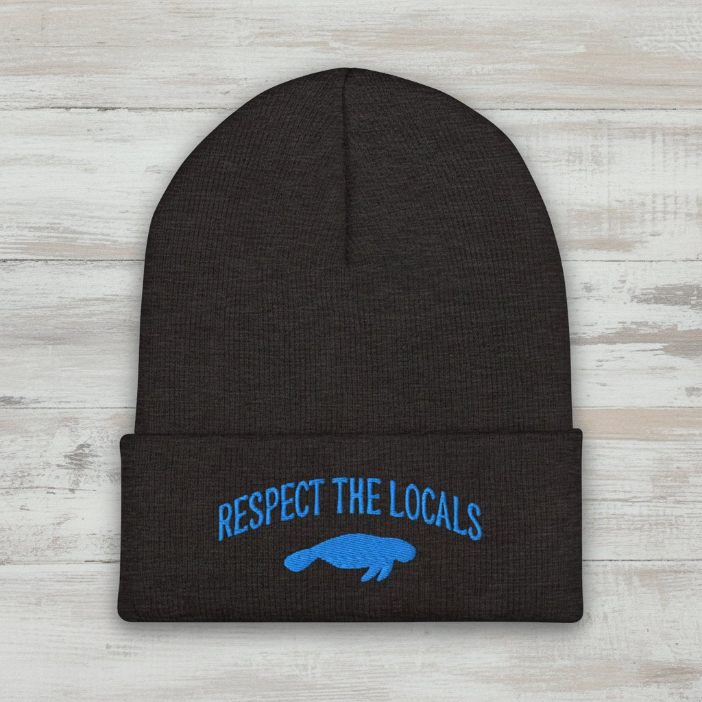 A dark charcoal gray heather knit beanie with a folded cuff, featuring blue embroidered text reading 'RESPECT THE LOCALS' and a blue manatee graphic on the front, set against a rustic wood background.