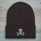 A dark charcoal gray knit beanie with a folded cuff, featuring an embroidered white skull and crossbones design on the front.