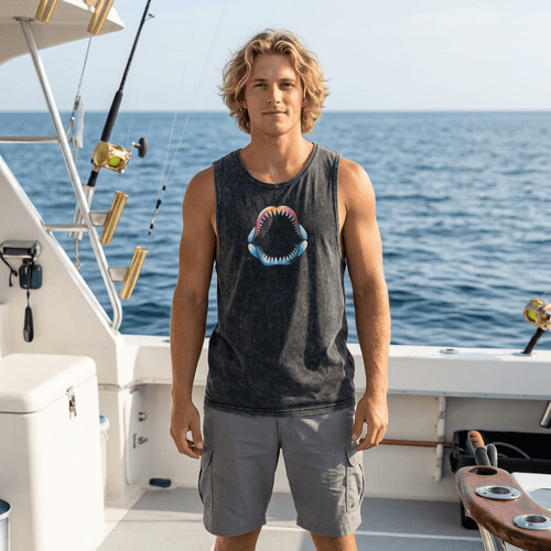 A man standing on a boat wearing a dark gray mineral-wash tank top with a colorful Shark Jaw graphic on the chest, paired with gray cargo shorts, with the ocean in the background.