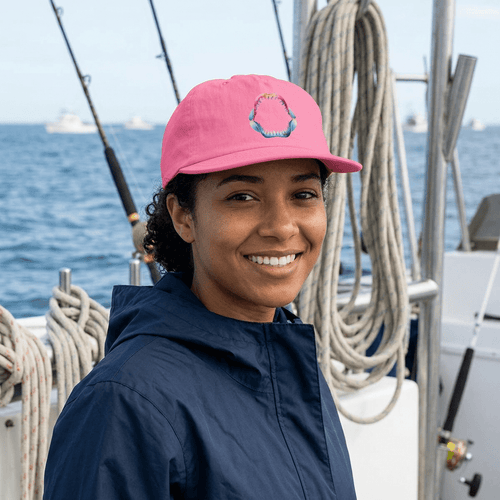 A smiling woman on a boat wearing a bright pink baseball cap with a small colorful shark jaw and a navy blue jacket, with ropes and blue water in the background.