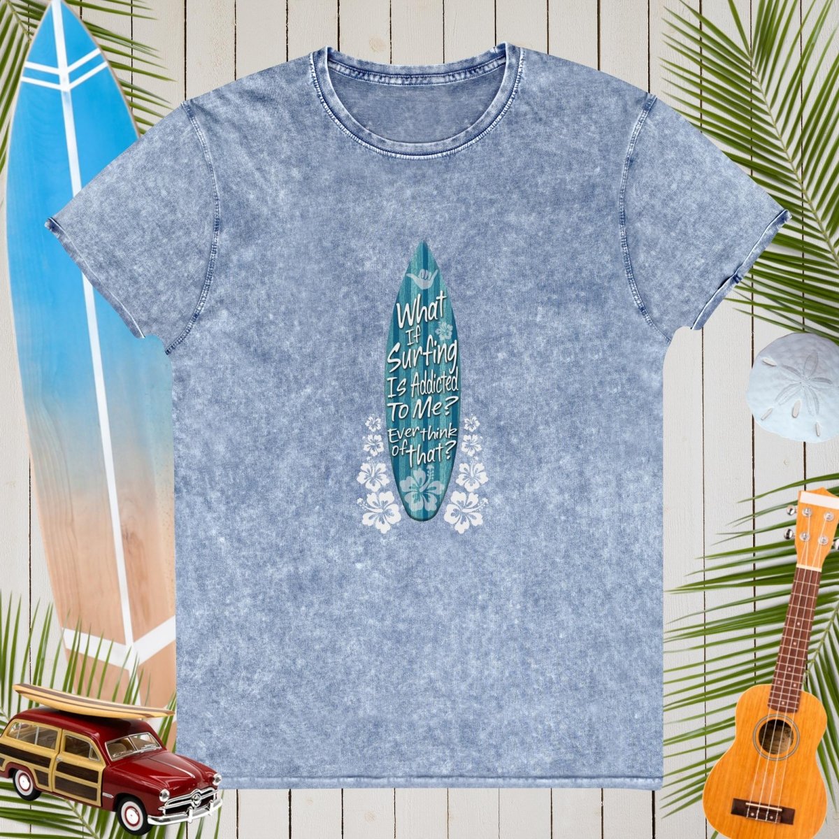 Blue t-shirt with a surfboard graphic and text on a wooden background with beach elements.
