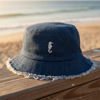 Navy blue denim bucket hat with frayed edge and seahorse embroidery on a beachside boardwalk