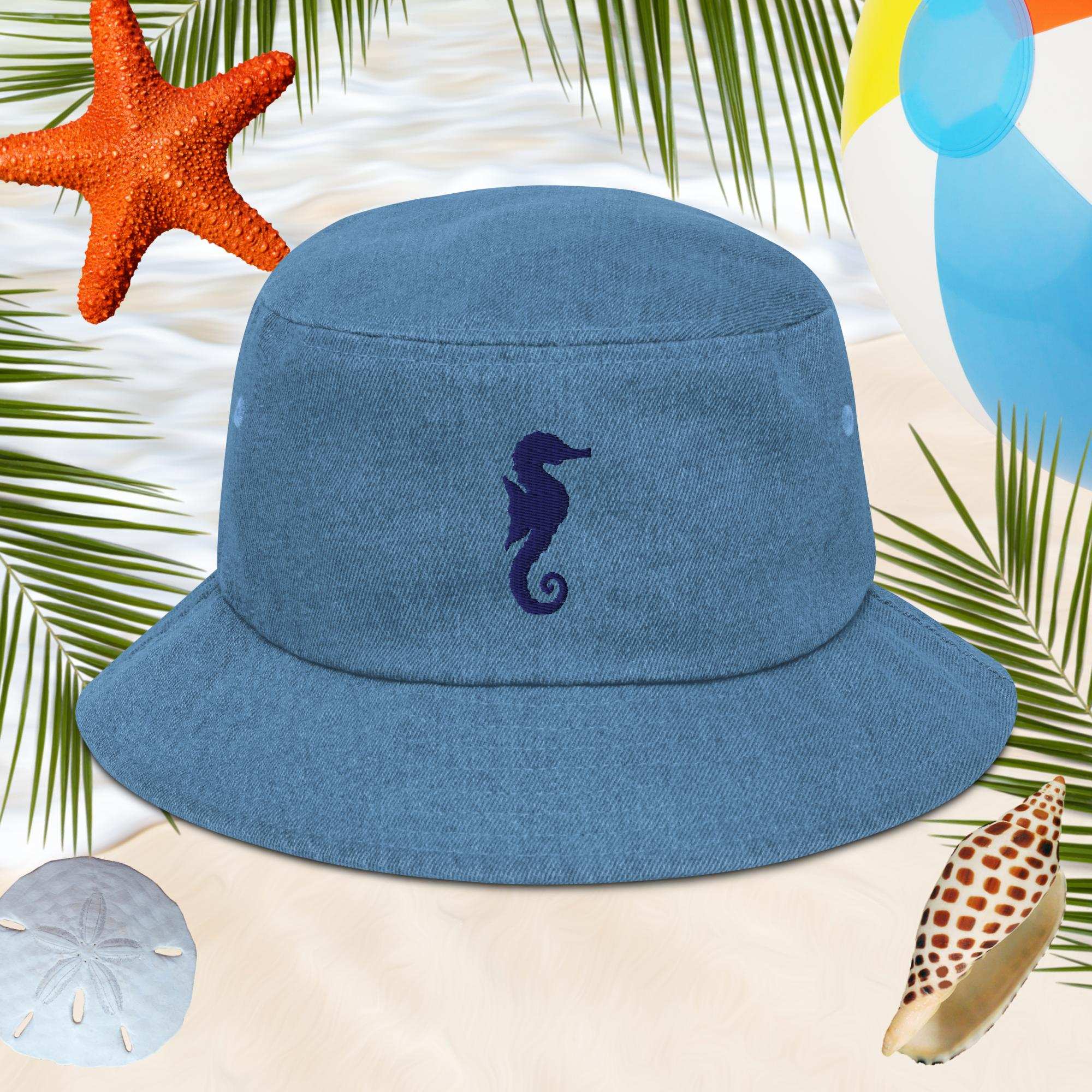 Blue bucket hat with an embroidered seahorse design on a beach-themed background with starfish, sand dollars, and shells.