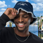 Man wearing a blue Embroidered Manatee bucket hat with text, smiling outdoors by water