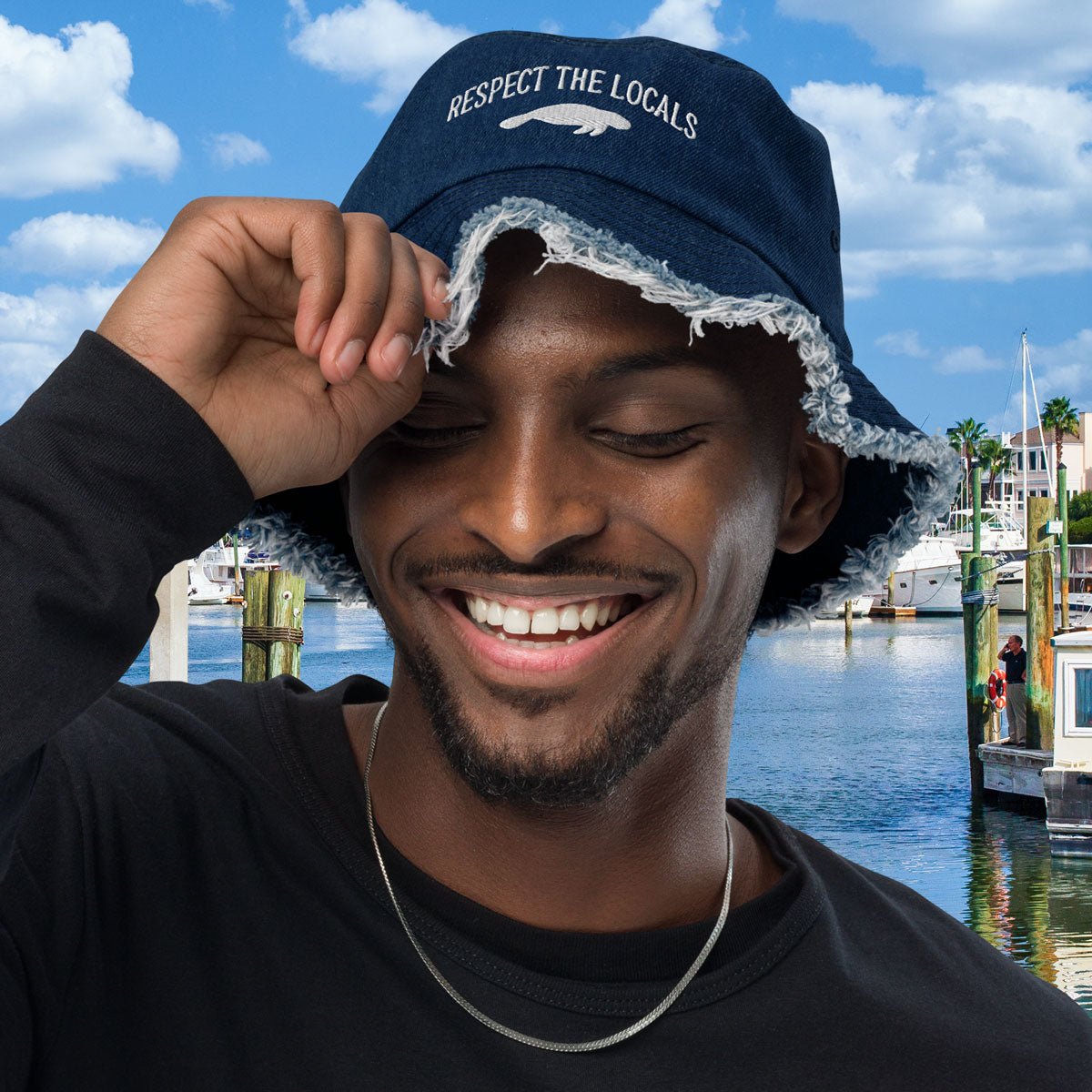 Man wearing a blue Embroidered Manatee bucket hat with text, smiling outdoors by water