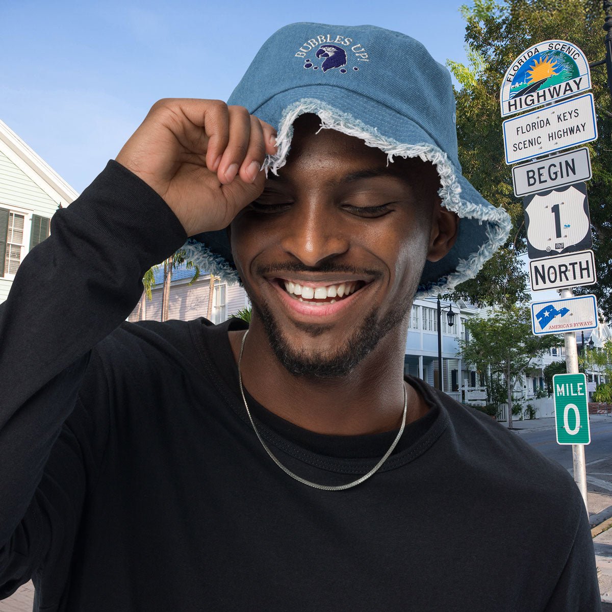 Man wearing a blue Bubbles Up Bucket Hat with a logo, smiling outdoors with scenic highway signs in the background.