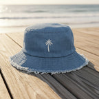 A light blue denim bucket hat with a white embroidered palm tree on the front, resting on a wooden deck with a blurred ocean background.