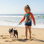 A young girl walking a Boston Terrier on a beach, wearing a long-sleeve dog swim shirt with orange sleeves and a colorful dog graphic on the front.