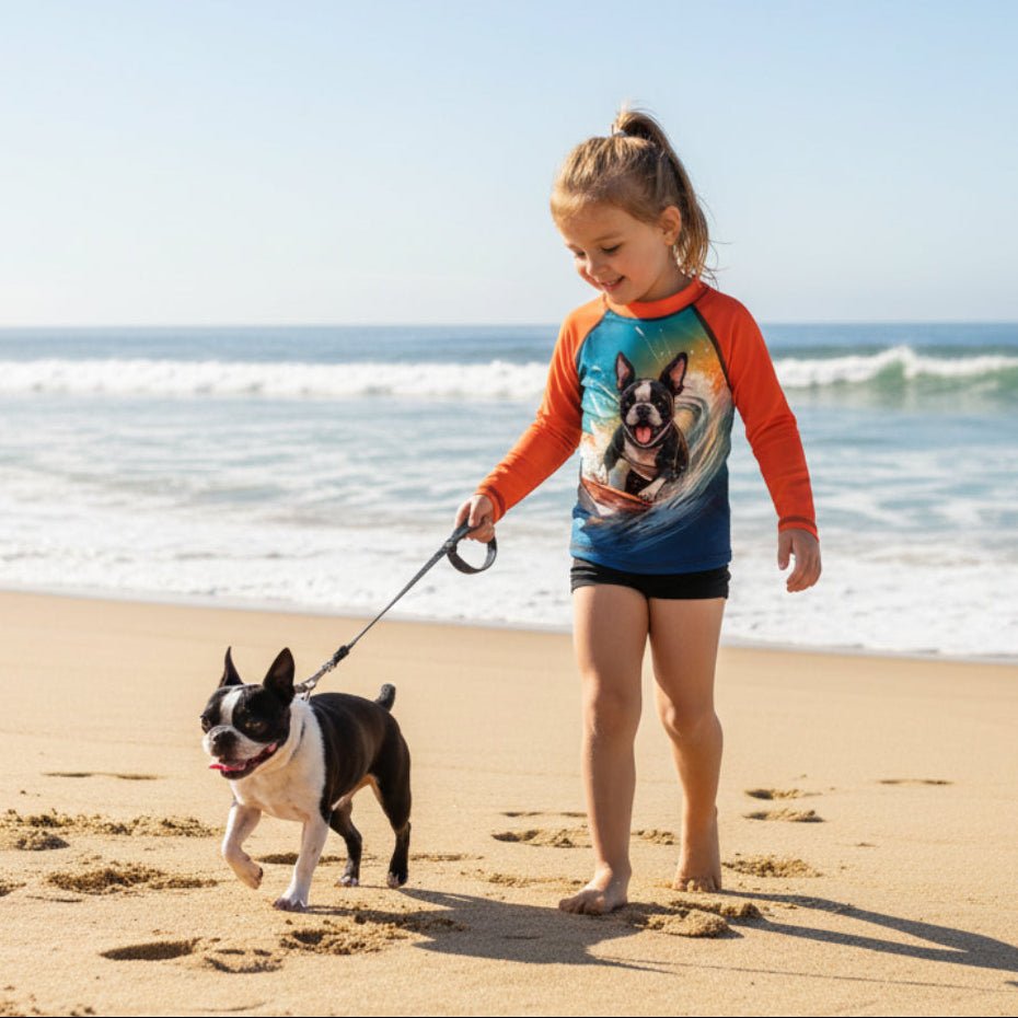 A young girl walking a Boston Terrier on a beach, wearing a long-sleeve dog swim shirt with orange sleeves and a colorful dog graphic on the front.