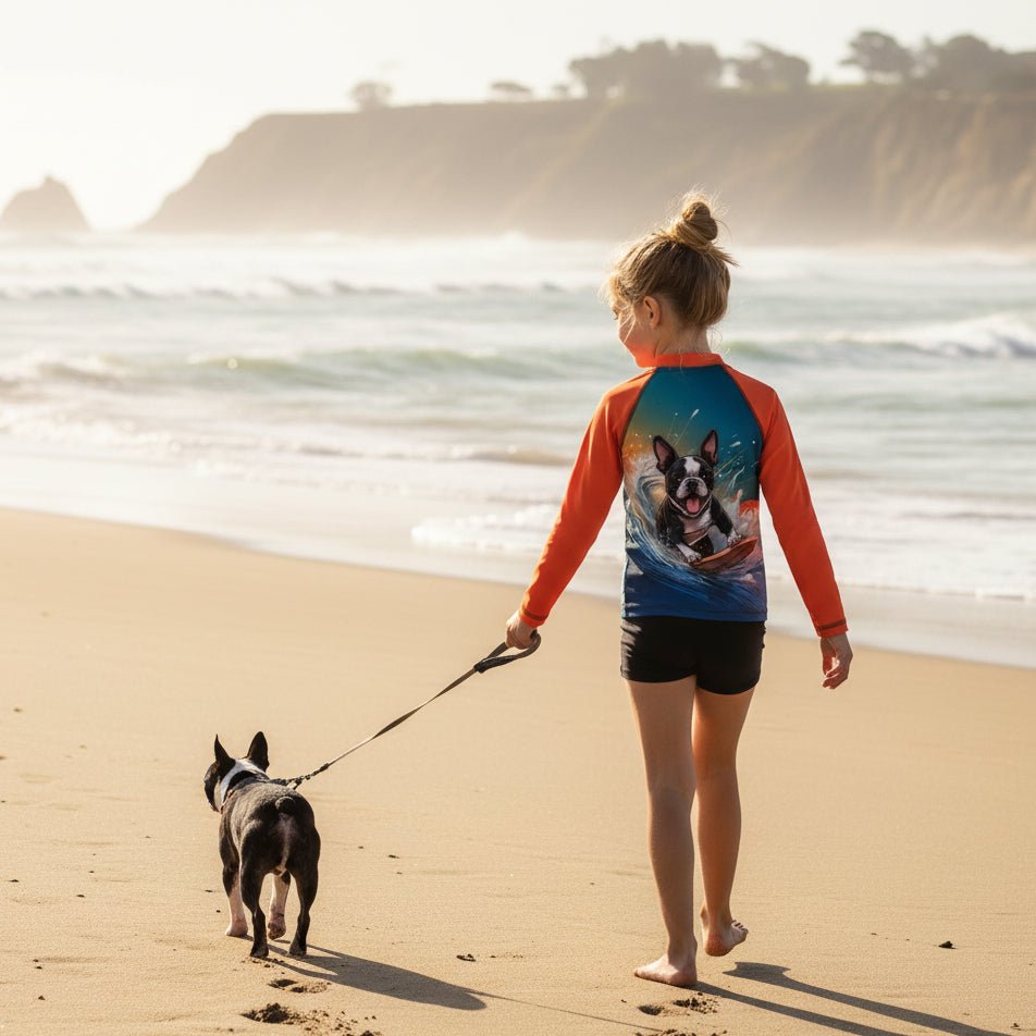 A child stands on a sandy beach facing the ocean, wearing a long-sleeve dog-print rash guard with bright orange sleeves and a blue graphic back, paired with black swim shorts. A small black dog walks beside them on a leash, with cliffs and sea in the background.