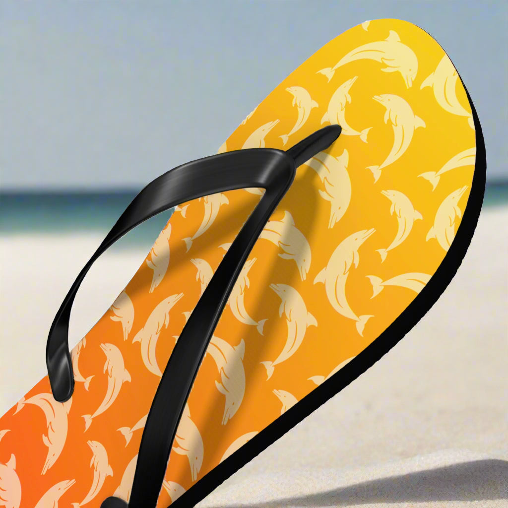 Flip-flop with dolphin pattern on a yellow and orange background sitting in the white sand of a pristine beach