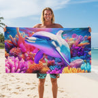 A smiling man stands on a sandy beach holding up a large rectangular towel or blanket printed with a colorful underwater scene featuring a dolphin swimming among bright coral reefs. The beach and ocean are visible in the background.