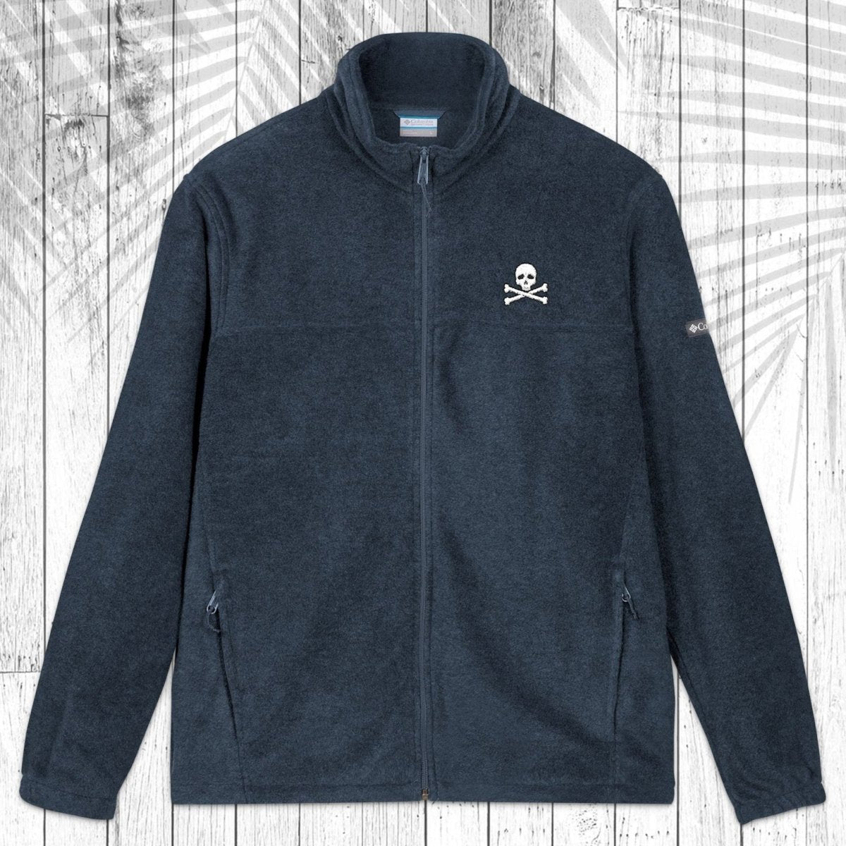A dark navy blue fleece zip-up jacket with a small white skull and crossbones embroidered on the left chest, displayed flat against a white wood plank background with a palm leaf overlay.