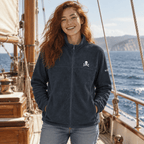 A woman with red hair stands on a wooden sailing ship deck wearing a dark navy fleece zip-up jacket with a small white skull and crossbones logo on the left chest, paired with light blue jeans. The background shows the blue ocean and distant cliffs.