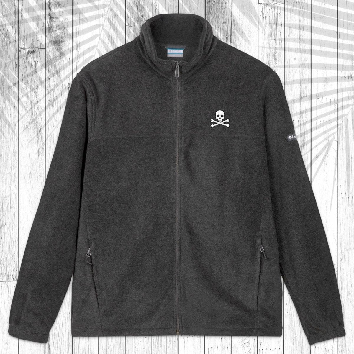 A dark gray fleece zip-up jacket with a stand collar, shown flat against a white wood plank background with a palm leaf pattern overlay. The jacket features a small white circular logo on the left chest and a small white logo on the right sleeve.