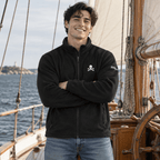 A smiling man wearing a black fleece zip-up jacket with a small white skull and crossbones logo on the left chest, standing on a wooden sailing ship with the sea in the background.