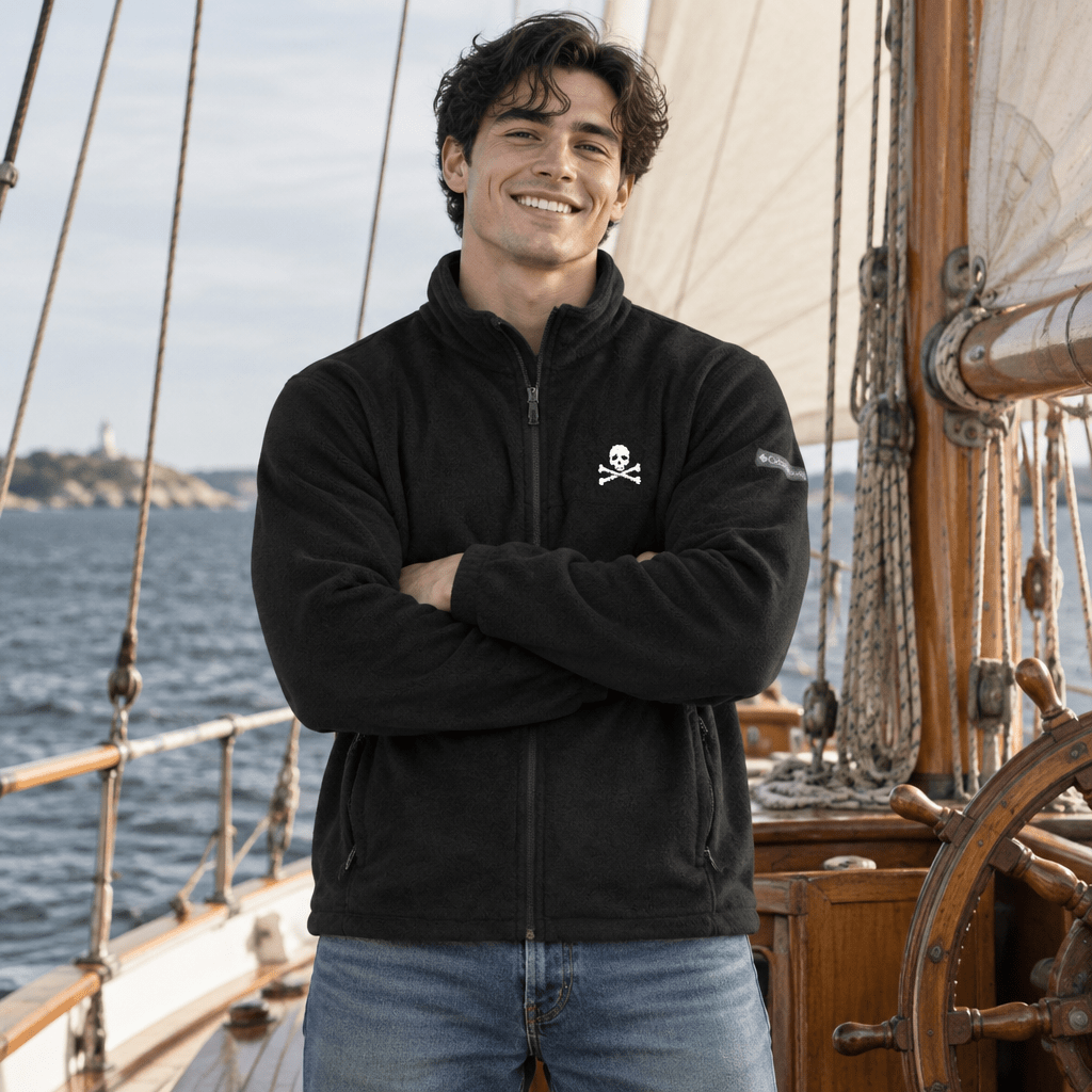 A smiling man wearing a black fleece zip-up jacket with a small white skull and crossbones logo on the left chest, standing on a wooden sailing ship with the sea in the background.