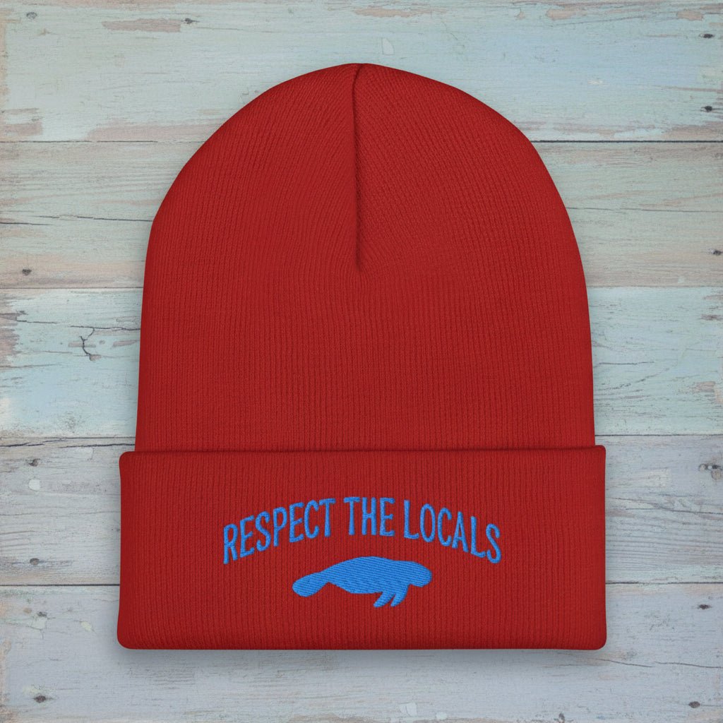 A red ribbed knit beanie with a folded cuff featuring blue embroidered text reading 'RESPECT THE LOCALS' above a blue manatee graphic, shown on a faded nautical wood background.