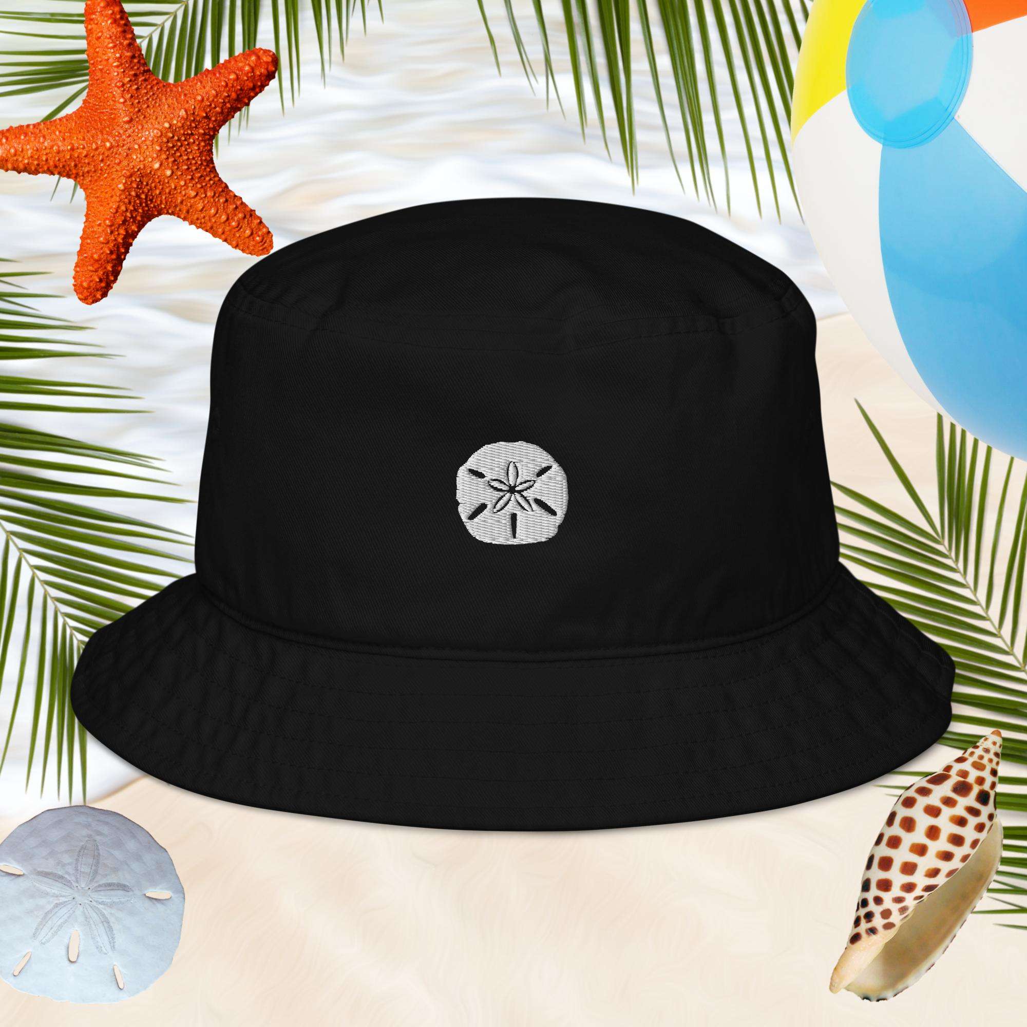 Black bucket hat with embroidered sand dollar design on a beach-themed background with starfish, shells, and palm leaves.