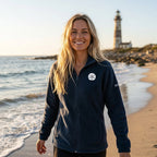 A woman stands on a beach wearing a dark navy fleece zip-up jacket with a white sand dollar embroidered on the left chest, paired with black pants.