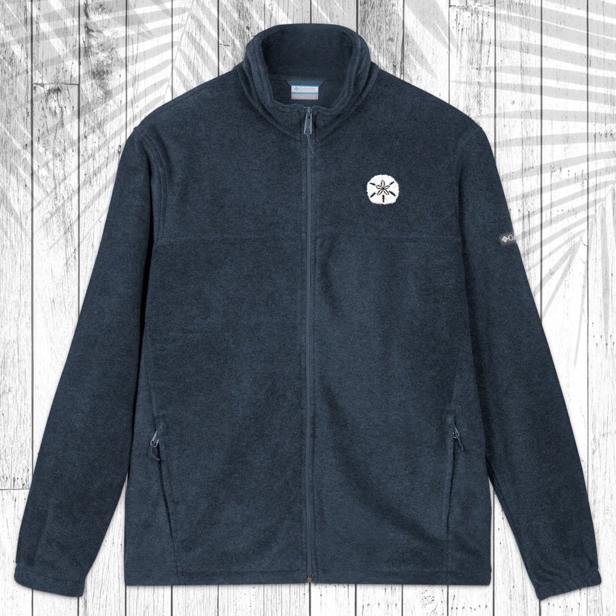 A dark navy blue fleece jacket with a full-zip front, stand-up collar, and zippered hand pockets, laid flat on a white wood plank background with a palm leaf pattern overlay.