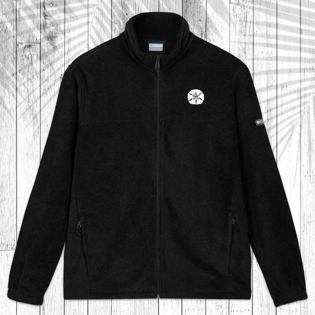 A black fleece zip-up jacket with a stand collar, shown flat against a white wood plank background with a leaf pattern overlay. It features a small white embroidered sand dollar on the left chest and a small white logo on the right sleeve.