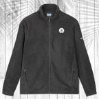 A dark gray fleece zip-up jacket with a stand collar, shown flat against a white wood plank background with a palm leaf pattern overlay. The jacket features a small white sand dollar embroidered on the left chest and a small white Columbia logo on the right sleeve.