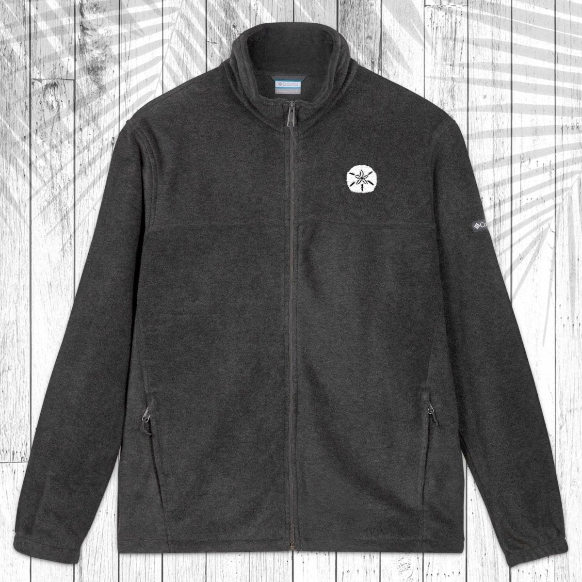 A dark gray fleece zip-up jacket with a stand collar, shown flat against a white wood plank background with a palm leaf pattern overlay. The jacket features a small white sand dollar embroidered on the left chest and a small white Columbia logo on the right sleeve.
