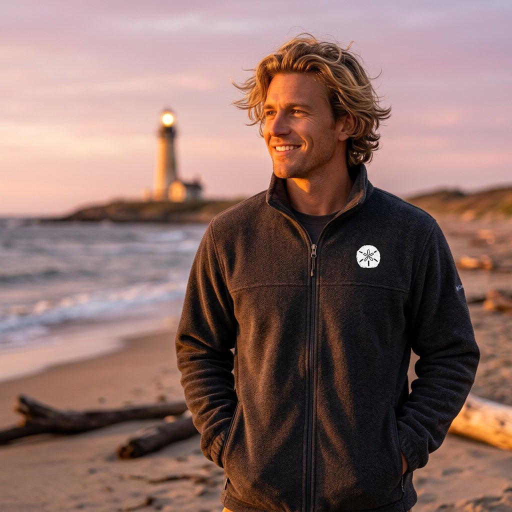 A man standing on a sandy beach wearing a dark charcoal gray zip-up fleece jacket with a white sand dollar embroidered on the left chest, paired with khaki pants.