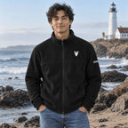 A man wearing a black fleece zip-up jacket with a white embroidered shark tooth stands on a rocky beach with the ocean and a lighthouse in the background.