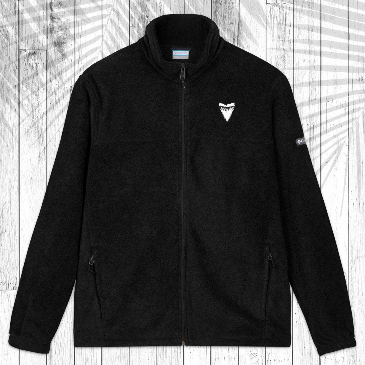A black fleece zip-up jacket with a stand collar, shown flat against a white wood plank background with a palm leaf shadows pattern overlay. The jacket features a small white shark's tooth on the left chest and a small white Columbia logo on the right sleeve.