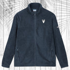 A dark gray fleece zip-up jacket with a stand collar, shown flat against a white wood plank background with a palm leaf pattern overlay. The jacket features a small white shark tooth embroidered on the left chest and a small white Columbia logo on the right sleeve.