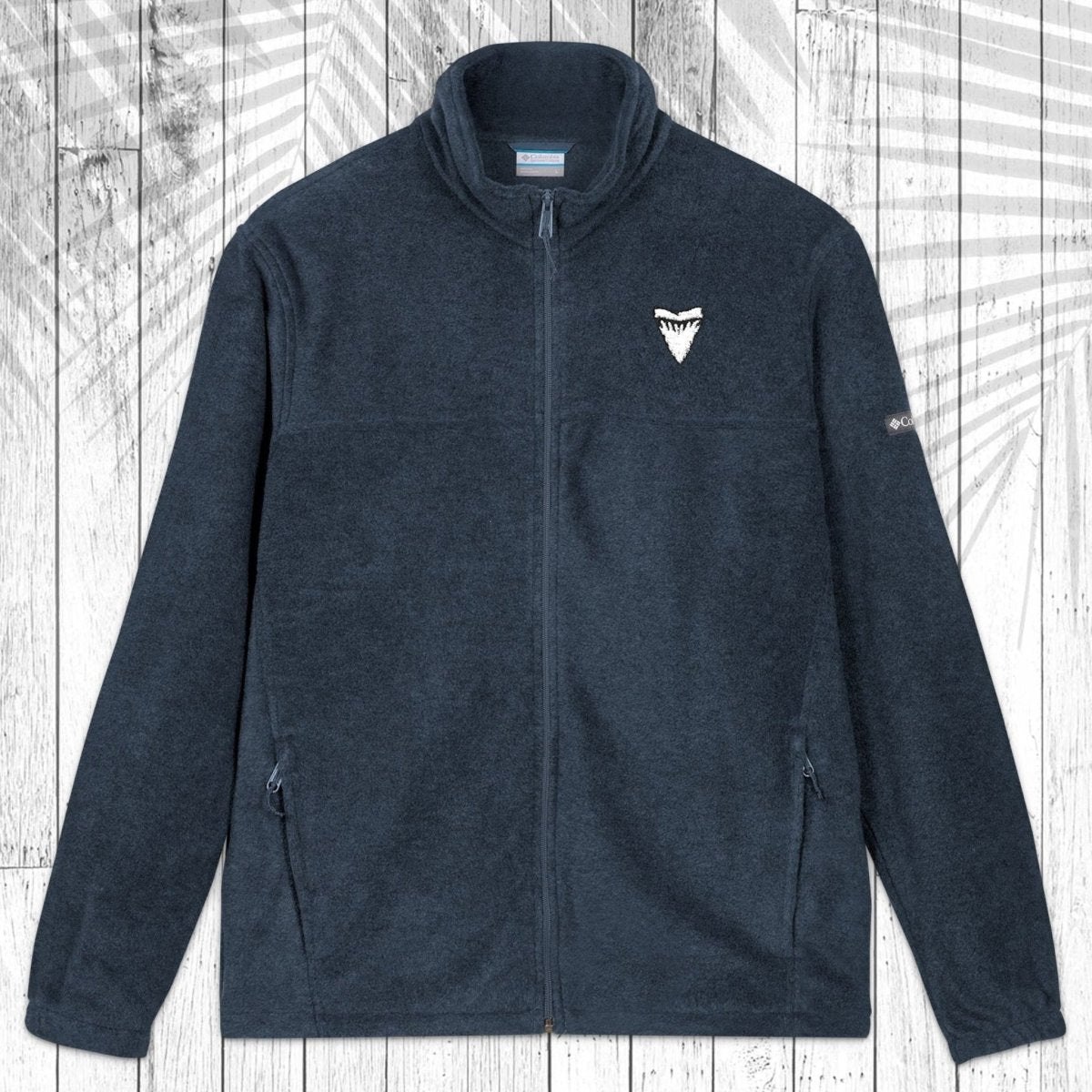 A dark gray fleece zip-up jacket with a stand collar, shown flat against a white wood plank background with a palm leaf pattern overlay. The jacket features a small white shark tooth embroidered on the left chest and a small white Columbia logo on the right sleeve.