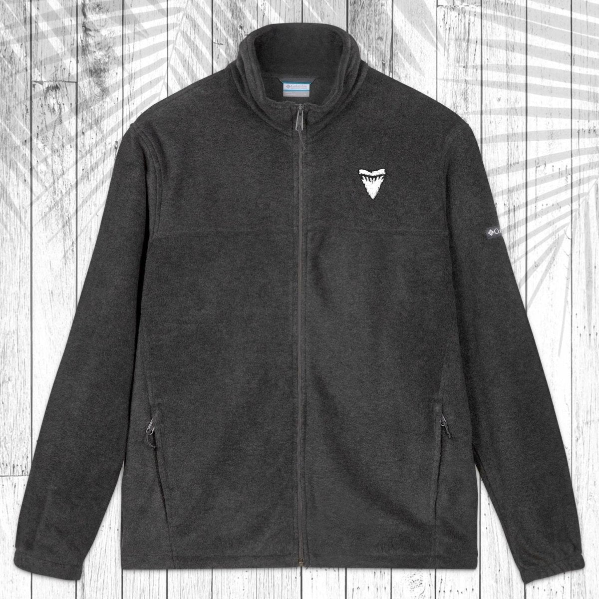 A dark gray fleece zip-up jacket with a stand collar, shown flat against a white wood plank background with a palm leaf pattern overlay. The jacket features a small white shark tooth embroidered on the left chest and a small white Columbia logo on the right sleeve.