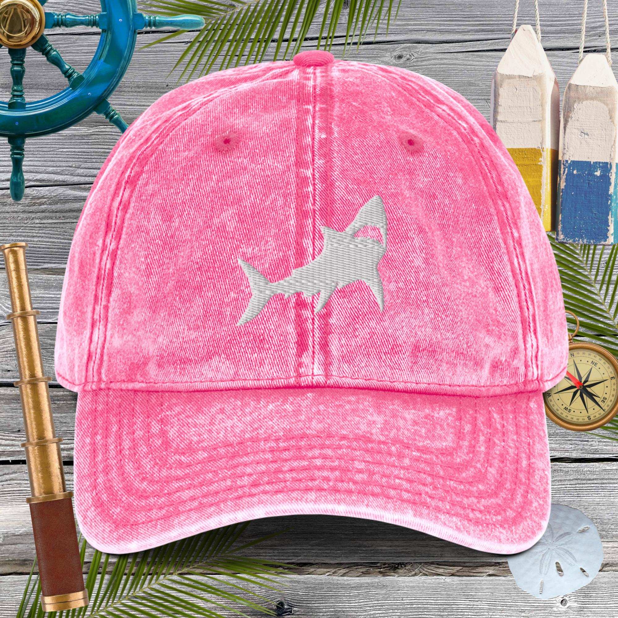 Pink cap with a shark design on a wooden background with nautical items.