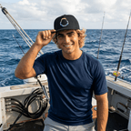 A smiling man on a boat wearing a navy blue short-sleeve shirt and a black baseball cap with a colorful circular graphic, with the ocean in the background.
