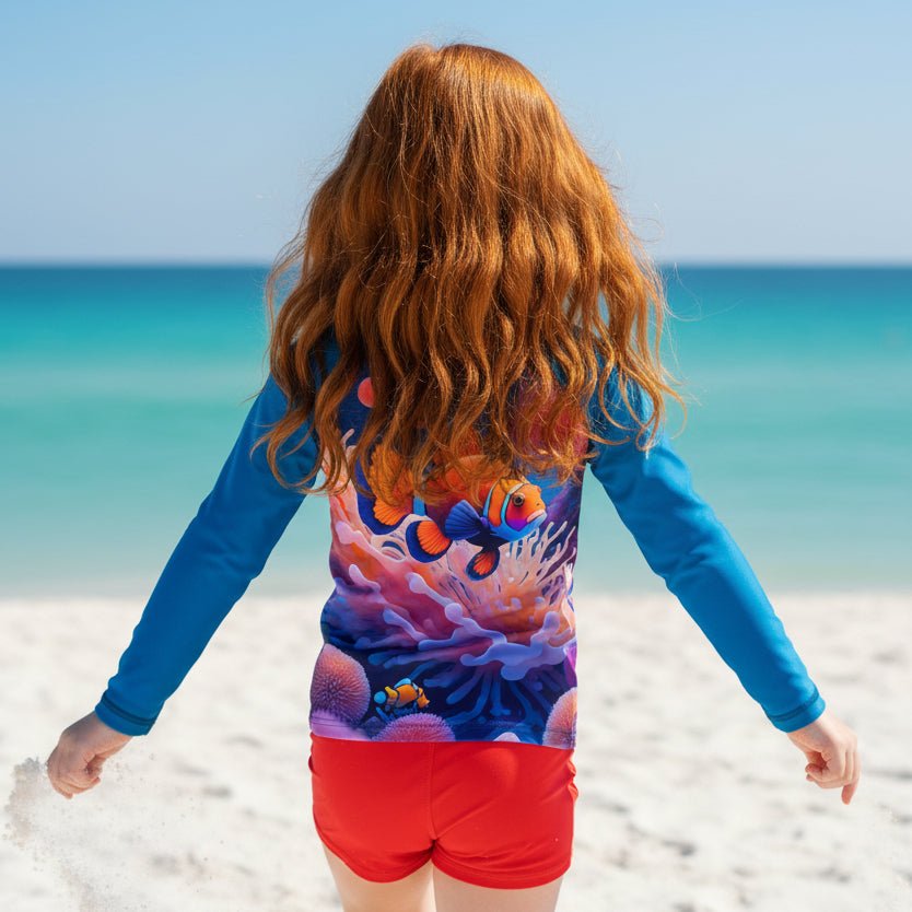 A long-sleeve raglan rash guard shirt featuring a vibrant underwater graphic print of a clownfish swimming among colorful coral reefs, worn by a little redhead girl on Fort Myers Beach