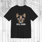 A black short-sleeve t-shirt with a graphic of a French Bulldog decorated with a colorful floral pattern and the text 'SHELL HOUND' printed below in white, displayed on a white wood plank background.